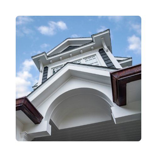 Versatex trim on a blue and white house, featuring white trim with a mid to dark brown wood-like section. The image is taken from below the front overhang, looking up at the sky.